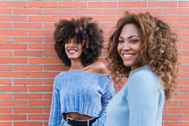 Amigos afroamericanos sonrientes con el pelo largo afro, maquillaje glamuroso y jersey azul posando en la pared de ladrillo. Belleza mujer de piel negra Africana Cara femenina étnica. Centrarse en el fondo - Foto, imagen
