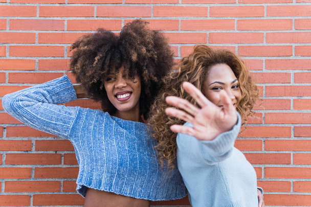 Amici neri positivi con lunghi capelli afro, trucco glamour e maglione blu che tira la mano verso la fotocamera. Ritratto di bellezza di ragazze africane naturali sorridenti nel muro di mattoni - Foto, immagini