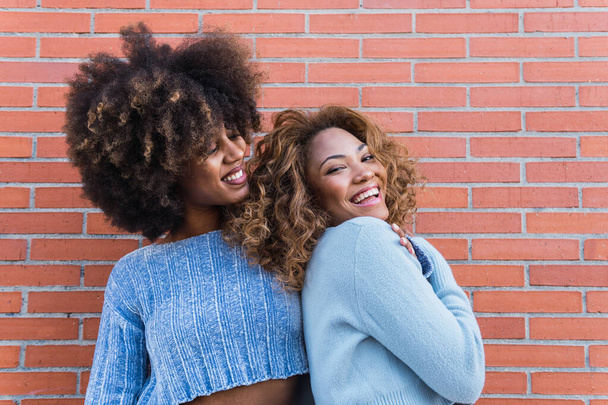 Smiling african american women with long afro hair, glamour makeup and blue shirt posing in brick wall. Beauty portrait of african natural girls having fun while hugging - Foto, Bild