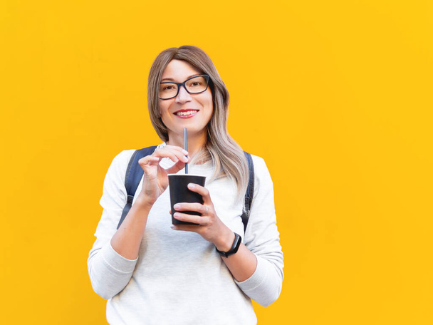 Wide smiling woman drinks coffee with black straw. Portrait of happy female with tasty cappuccino on monochrome yellow wall background. Bright and colorful backdrop. Student with coffee to go. - Foto, Imagen