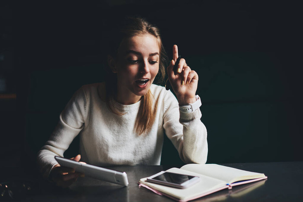 Young female freelancer in casual outfit with opened mouth and finger up sitting at table with opened notebook and tablet looking at smartphone screen - Foto, Imagem