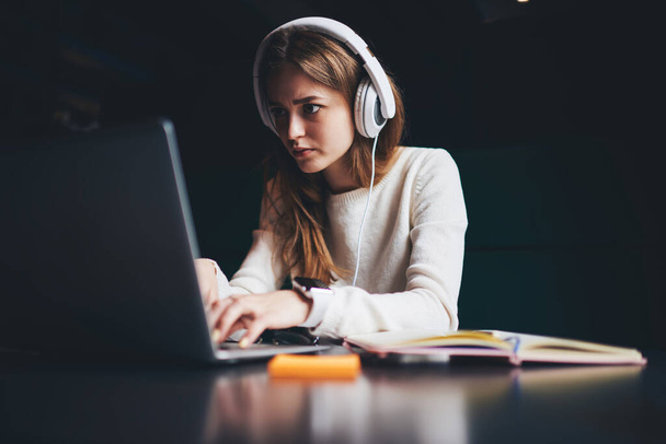 Concentrated female teleworker in casual outfit and headphones attentively looking at screen and typing while working on laptop remotely in creative workspace - Фото, изображение