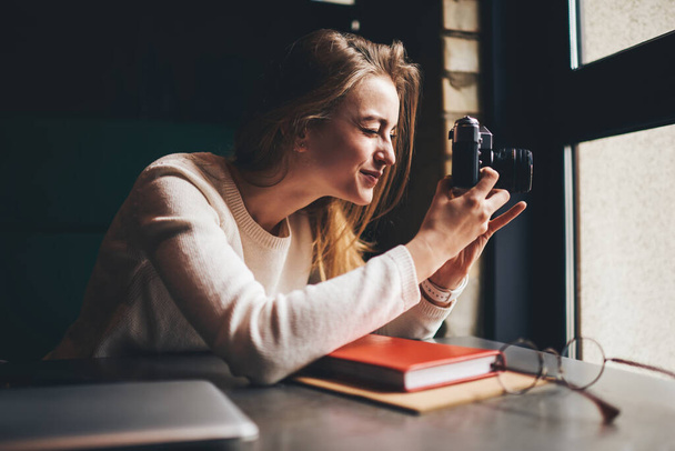 Side view of young female photographer with analog photo camera sitting at table with books and squinting eye while shooting through window - Photo, Image