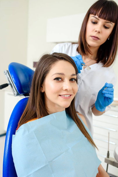 Mladá šťastná usměvavá pacientka na zubní klinice podstupuje stomatologické ošetření a dívá se do kamery. Zubař se zubním nářadím je vzadu. - Fotografie, Obrázek