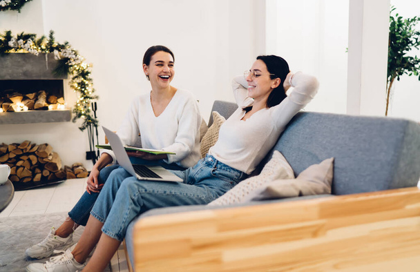 Positive women in casual clothes sitting on comfortable sofa and laughing while using laptop and discussing project in light cozy room - Photo, image