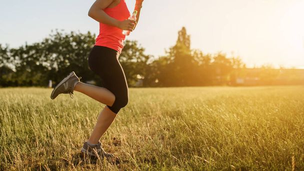 Spor yapan bir kadının bacaklarını kapayarak gün batımında doğanın ortasında çimlerin üzerinde koşuyor. Her yerde spor yap. - Fotoğraf, Görsel