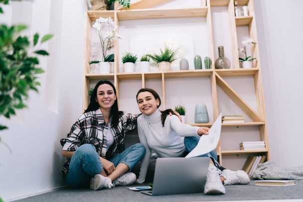 Positive female coworkers sitting close and smiling brightly while doing remote job together with laptop and documents in modern apartment - Photo, Image