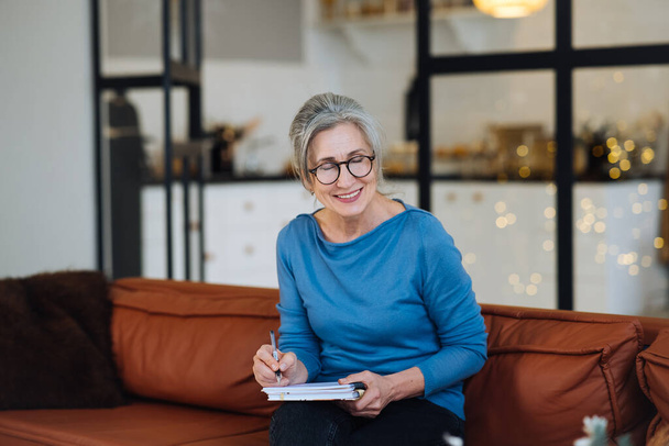 Feliz mujer mayor en gafas de escribir a cuaderno en casa - Foto, imagen