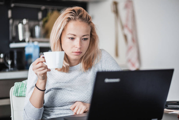 Female freelancer in her casual home clothing working remotly from her dining table in the morning. Home kitchen in the background. - Photo, Image