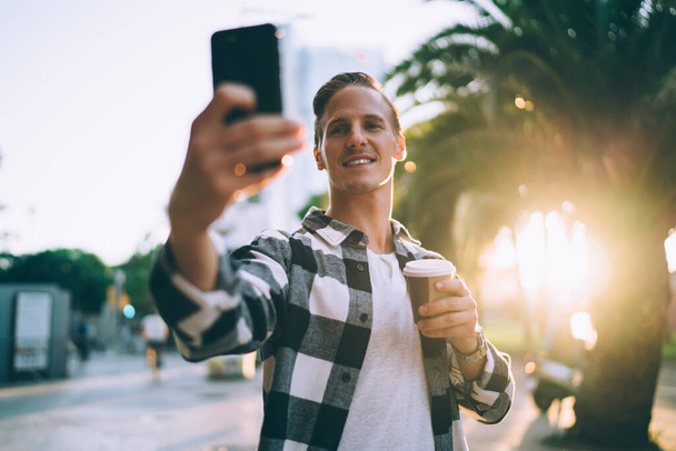 Positive young man generation z making picture of spending time in city holding coffee to go on sunny evening free time, handsome male influencer posing for selfie enjoying weekends on urban setting - Photo, Image