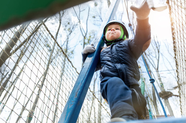 Κάτω POV άποψη γενναίο θάρρος μικρό παιδί αγόρι φορούν κράνος εξοπλισμού ασφαλείας απολαμβάνουν περνώντας εμπόδιο πορεία δάσος σχοινί περιπέτεια πάρκο την κρύα ημέρα του χειμώνα. Δραστηριότητα εκτός κατασκήνωσης αναψυχής - Φωτογραφία, εικόνα