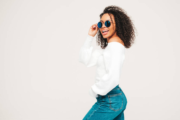 Beautiful black woman with afro curls hairstyle.Smiling model in sweater and trendy jeans clothes. Sexy carefree female posing on white background in studio. Tanned and cheerful. In sunglasses - Φωτογραφία, εικόνα