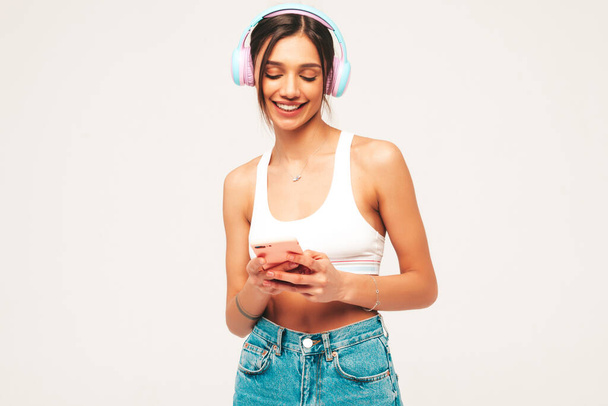 Beautiful smiling woman dressed in jeans clothes. Sexy carefree model listening music in wireless headphones. Adorable and positive female posing on grey background in studio. Holding smartphone - Fotoğraf, Görsel