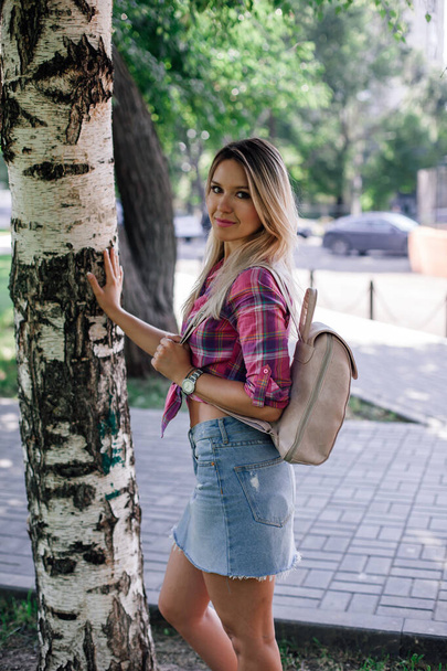 Adult shining blonde woman with face looking into camera standing in park near birch wearing checkered shirt and jeans skirt having backpack on in daytime. Adorable background full of green - Photo, Image