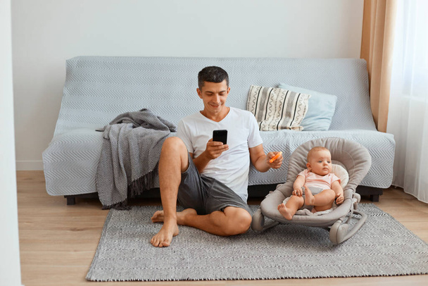 Concentrated young adult Caucasian man wearing white t shirt and short sitting on the floor with infant daughter in rocking chair, using mobile phone and playing with his toddler kid. - Foto, Imagen