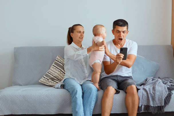 Indoor shot of young parents with their kid sitting on cough in light room, man holding cell phone, being shocked to win, betting, man screaming with amazement, woman giving baby to father. - Fotó, kép