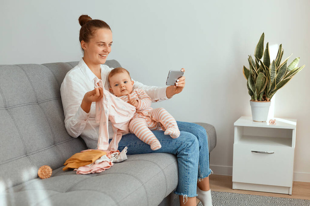 Indoor shot of smiling woman blogger shooting video for her mom's vlog, showing new kid's clothing to her followers while broadcasting livestream, expressing positive emotions. - Фото, зображення