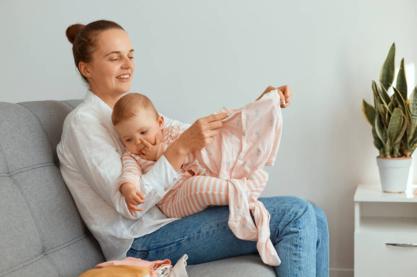 Woman in casual style clothes have fun with cute baby girl, holding infant daughter, admires new clothing for toddler child, sitting on sofa in living room. - Foto, immagini