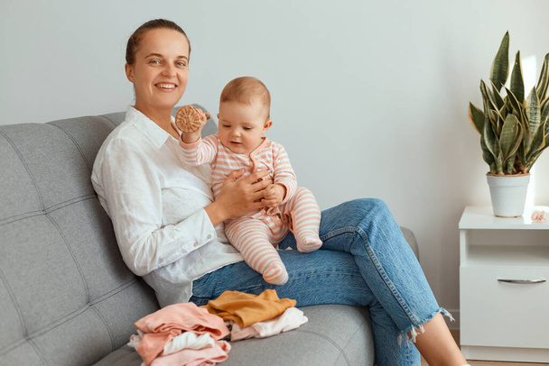 Image of smiling mother sitting on cough with her infant daughter and playing with toy, looking at camera with positive happy expression, happy childhood and motherhood. - Foto, imagen