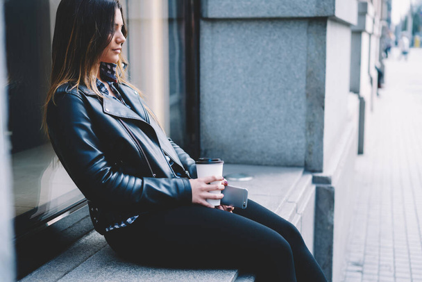 Side view of stylish female with paper cup of hot drink and mobile phone sitting on concrete windowsill on street - Foto, afbeelding