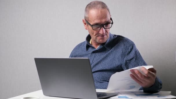 Älterer Mann checkt Zeichnungen neben Laptop am weißen Bürotisch - Filmmaterial, Video