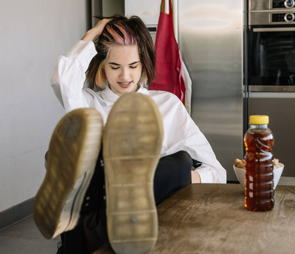 front view of a girl sitting with her feet on the kitchen table next to a bottle of soda, horizontal view - Photo, Image