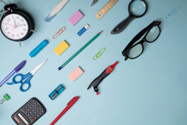 Paper stationery. School supplies, diverse pens, clips, markers, pencils, scissors, flash drive, eyeglasses, magnifying glass, and alarm clock on a blue background - Photo, Image