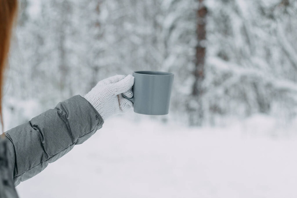 A hand in a white knitted woolen mitt and a gray jacket holds a gray mug against the background of a beautiful snow-covered forest. - Foto, afbeelding