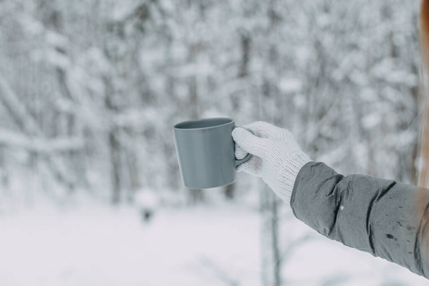 A hand in a white knitted woolen mitt and a gray jacket holds a gray mug against the background of a beautiful snow-covered forest. - Foto, Imagem