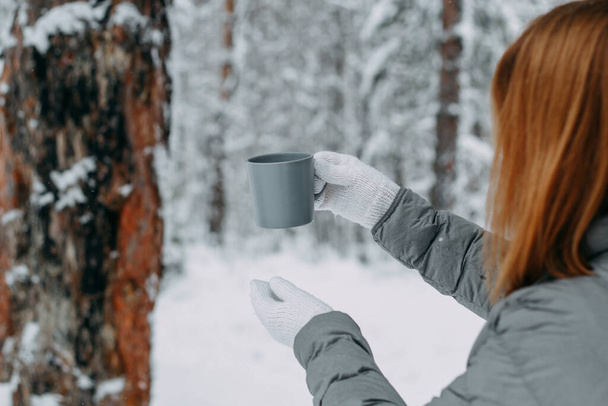 A beautiful girl holds out a mug with a hot drink to warm up. Warm and cozy in the cold snowy forest in winter.selective focus. - Фото, зображення