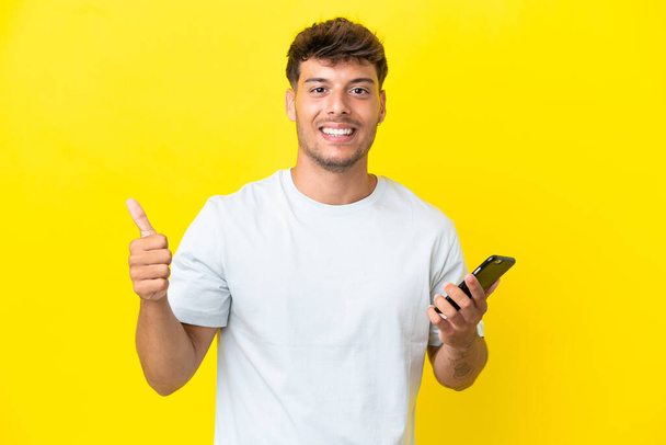 Young caucasian handsome man isolated on yellow background using mobile phone while doing thumbs up - Φωτογραφία, εικόνα
