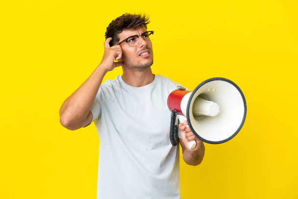 Young caucasian handsome man isolated on yellow background holding a megaphone and having doubts - Zdjęcie, obraz