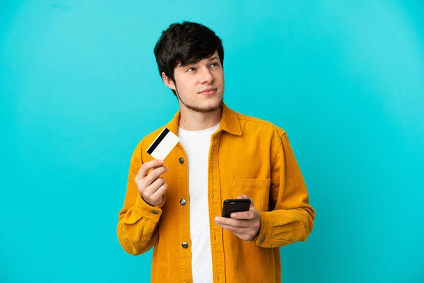 Młody Rosjanin odizolowany na niebieskim tle kupuje za pomocą telefonu komórkowego z karty kredytowej podczas myślenia - Zdjęcie, obraz