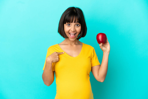 Kobieta w ciąży trzyma jabłko odizolowane na niebieskim tle z zaskakującym wyrazem twarzy - Zdjęcie, obraz