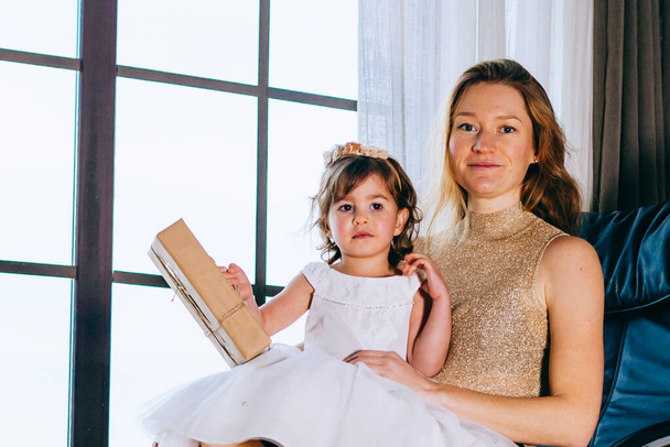 Niña en vestido blanco con su amorosa madre sentada en casa cerca de la maceta - Foto, Imagen