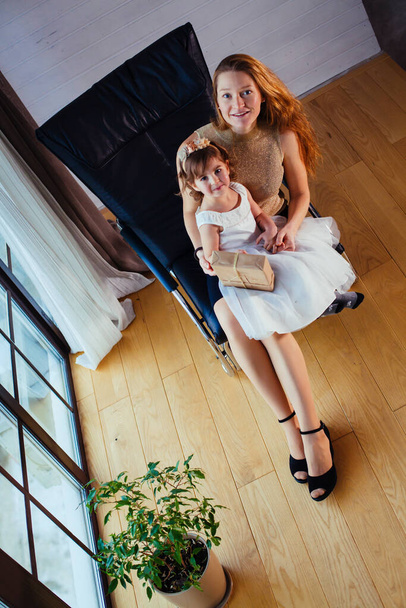 Маленькая девочка в белом платье с любящей матерью сидит дома рядом с цветочным горшком - Фото, изображение