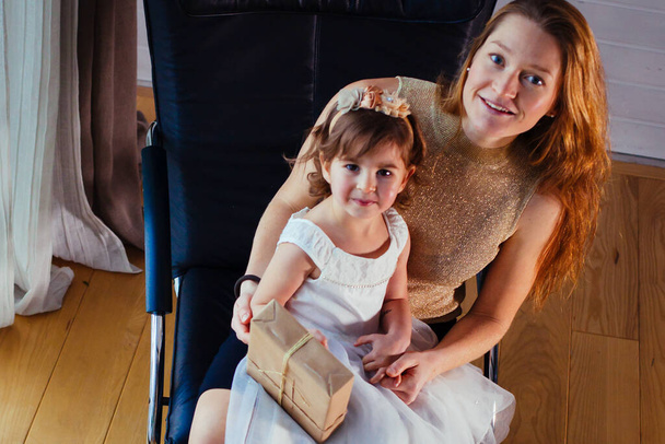 Little girl in white dress with her loving mother sitting at home  near the flower pot - Foto, Imagem