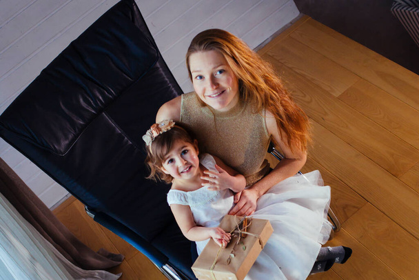 Niña en vestido blanco con su amorosa madre sentada en casa cerca de la maceta - Foto, imagen