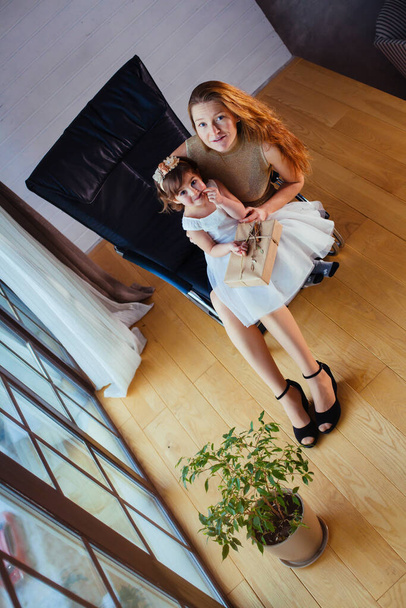 Niña en vestido blanco con su amorosa madre sentada en casa cerca de la maceta - Foto, Imagen