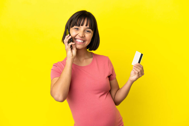 Joven mujer embarazada de raza mixta aislada sobre fondo amarillo manteniendo una conversación con el teléfono móvil y sosteniendo una tarjeta de crédito - Foto, Imagen