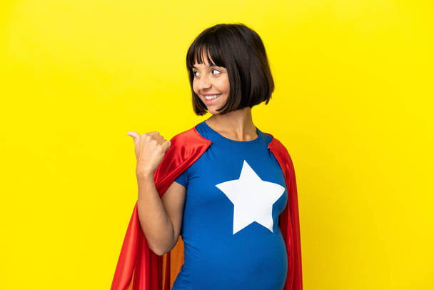 Супергерой беременная женщина изолированы на желтом фоне указывая в сторону, чтобы представить продукт - Фото, изображение