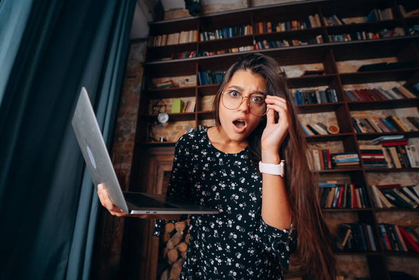 Jonge vrouw in bril kijkt geschokt na het controleren van e-mail op laptop - Foto, afbeelding