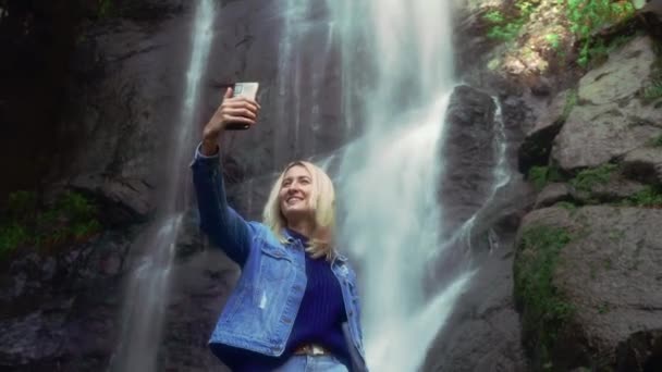 nainen omistaa älypuhelimen ja ottaa kuvia itsestään seisoo lähellä vesiputous - Materiaali, video