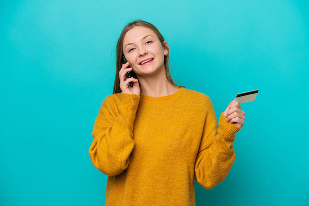 Fiatal orosz nő elszigetelt kék háttér tartja a beszélgetést a mobiltelefon és a kezében egy hitelkártya - Fotó, kép