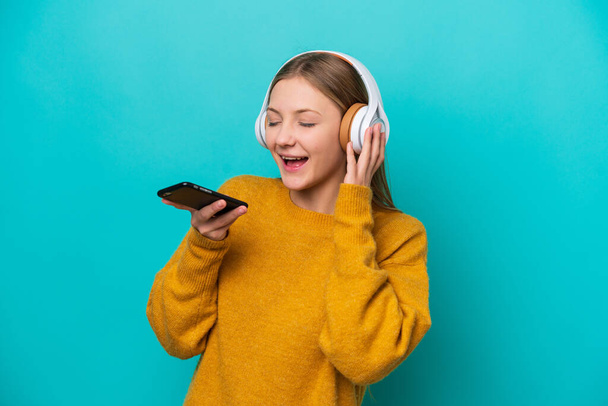 Молода російська жінка, ізольована на синьому фоні, слухає музику мобільним телефоном і співає - Фото, зображення