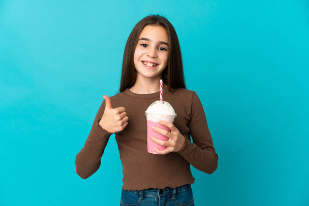 Маленькая девочка с клубничным молочным коктейлем изолирована на синем фоне, показывая большой палец вверх. - Фото, изображение