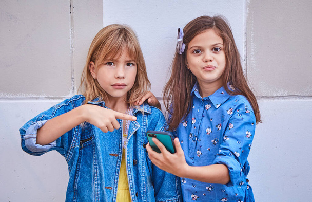 Niedliche zwei Freundinnen eines 9-jährigen Mädchens stehen zusammen an der frischen Luft und schauen auf das Telefon und zeigen mit der Hand auf das Telefon. Das Konzept der Freundschaft, Beziehungen von Jugendlichen - Foto, Bild