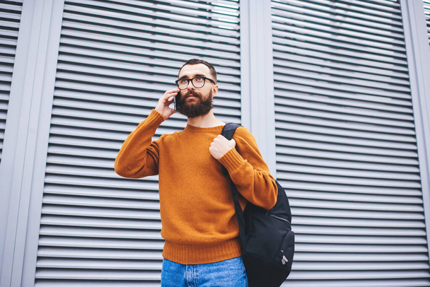 From below of bearded male in casual clothes with eyeglasses looking away making phone call while walking on city street - 写真・画像