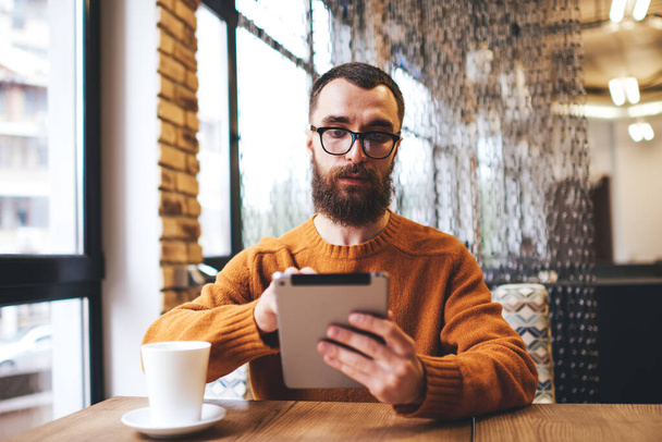 Unshaven male in casual clothes with eyeglasses sitting at table with cup of hot drink typing on laptop having rest in cafe - Foto, Imagen