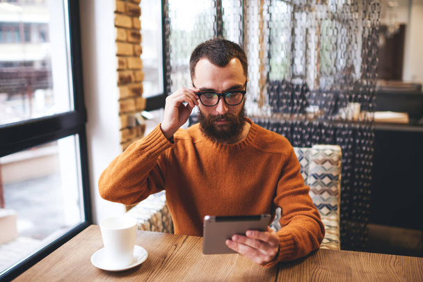 Concentrated bearded male sitting at table with cup of coffee touching eyeglasses looking at screen of tablet checking social network during break - Фото, зображення